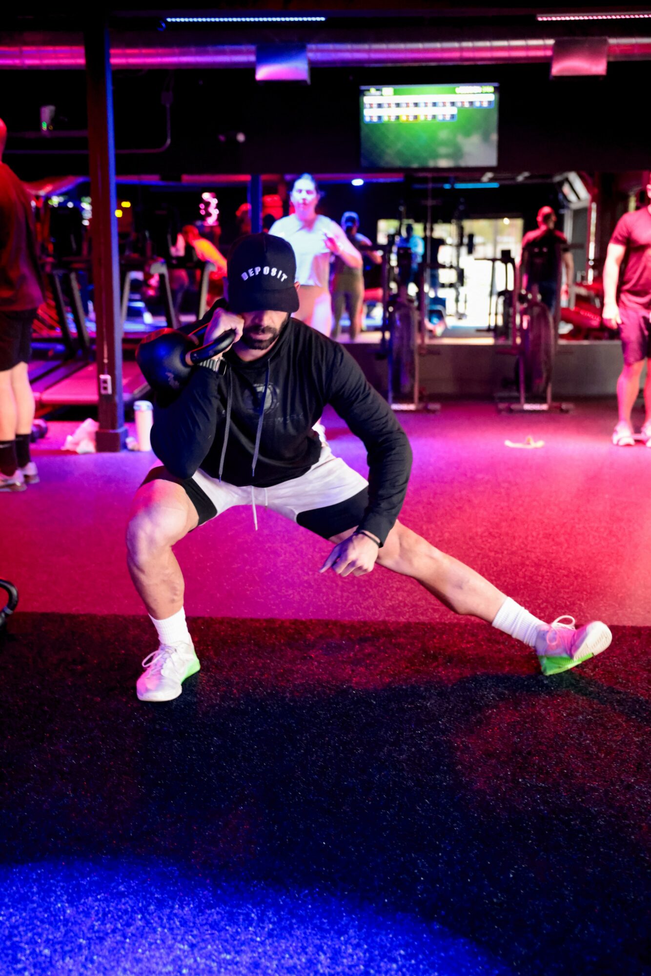 Person in gym with colorful lighting, stretching in front of mirrors, wearing black hoodie, shorts, and cap.