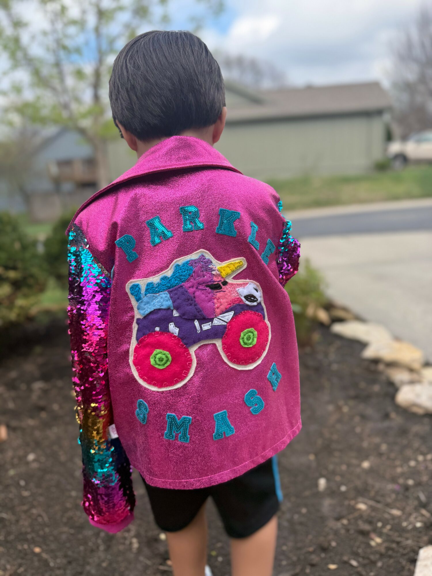 Child wearing a pink jacket with colorful sequins and a monster truck design, standing outdoors on a sidewalk.