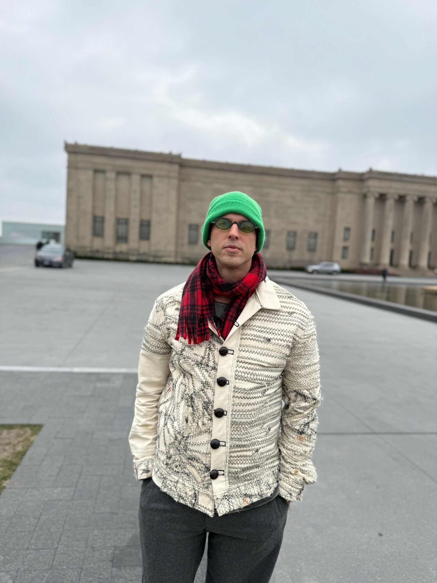 Person wearing a green beanie, red scarf, and patterned jacket standing outdoors in a parking lot with a large building in the background.