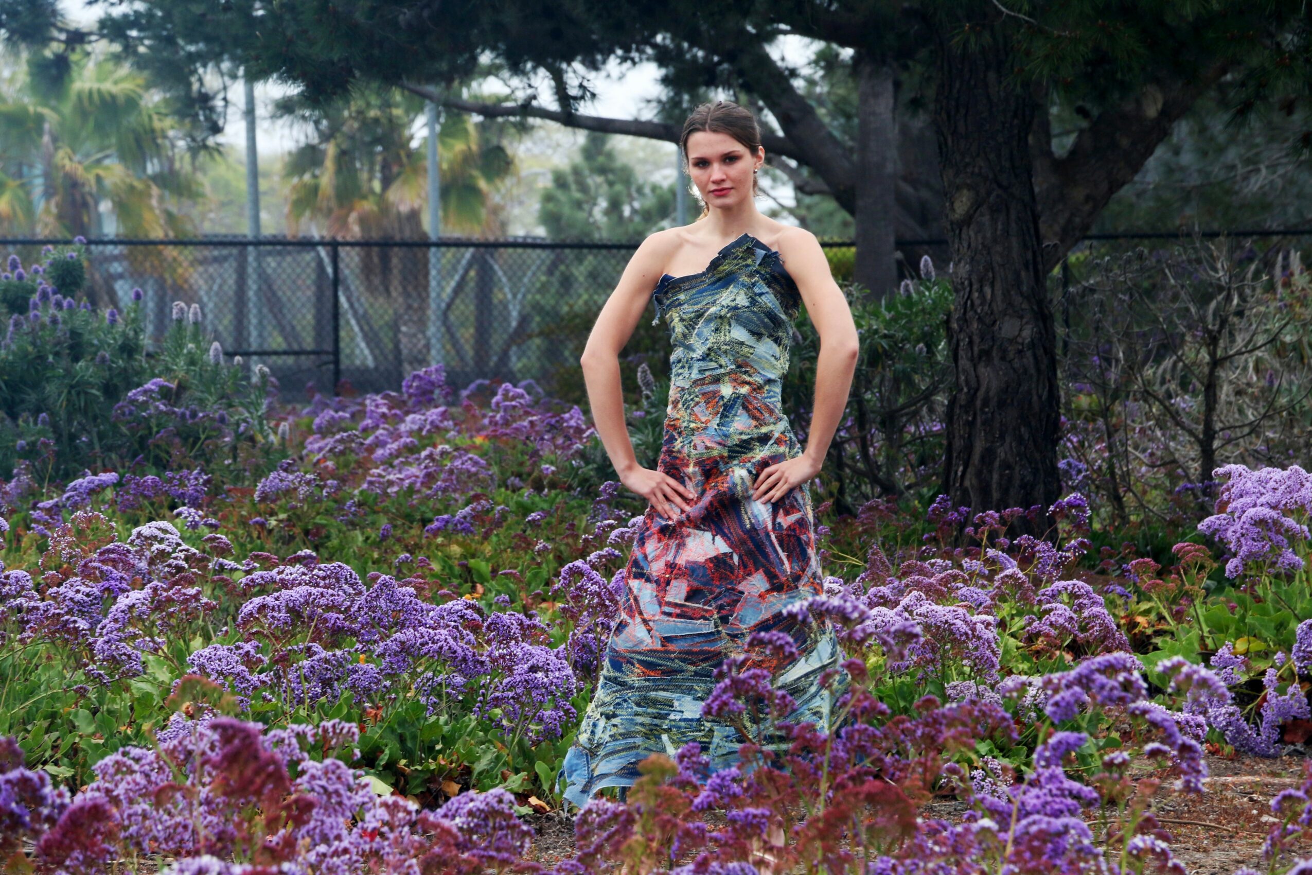 Woman standing in a garden with purple flowers, wearing a patterned dress, with trees and a fence in the background.