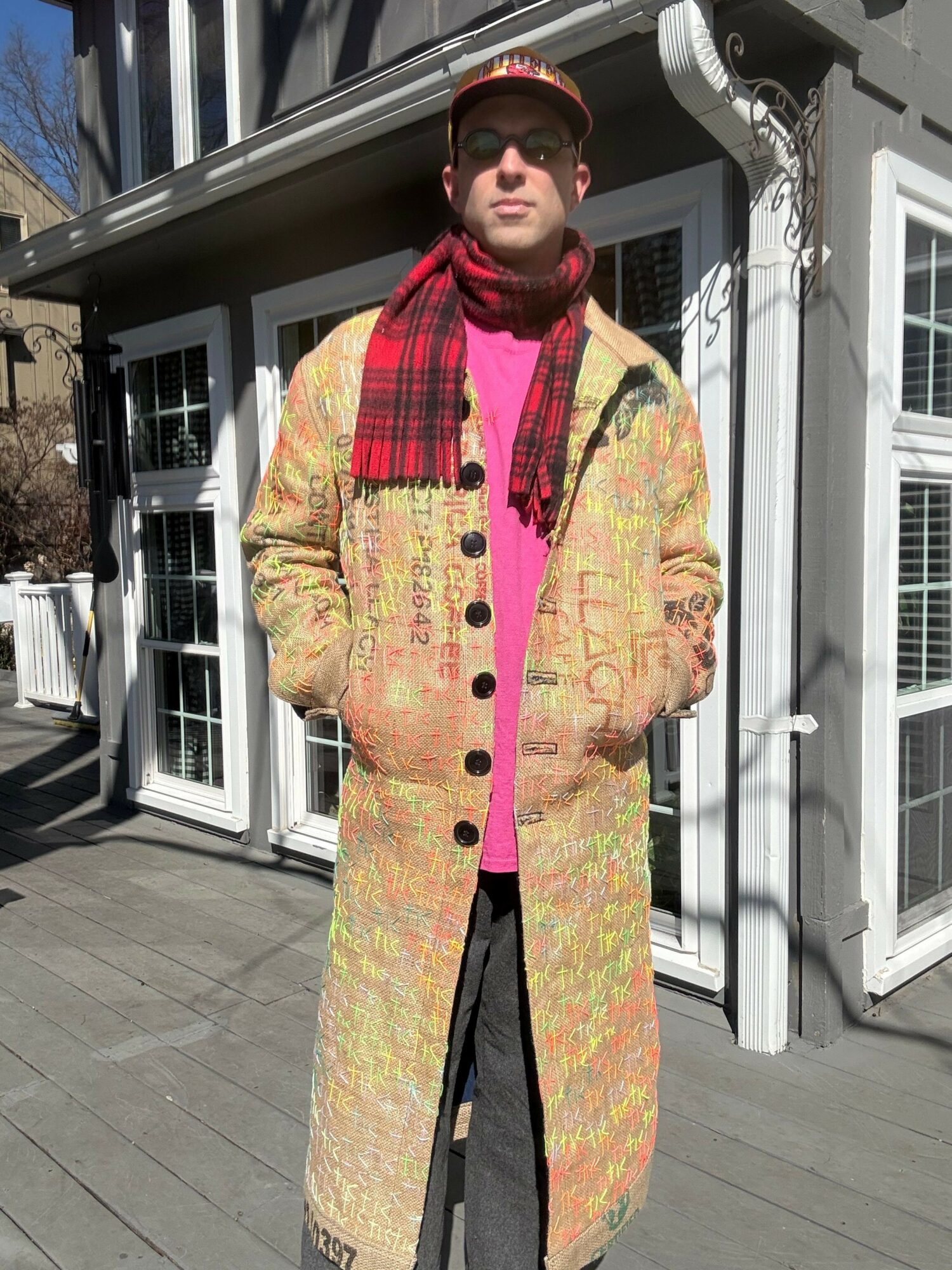 Person standing outdoors on a wooden deck, wearing a colorful patterned suit and a red cap, with a building in the background.