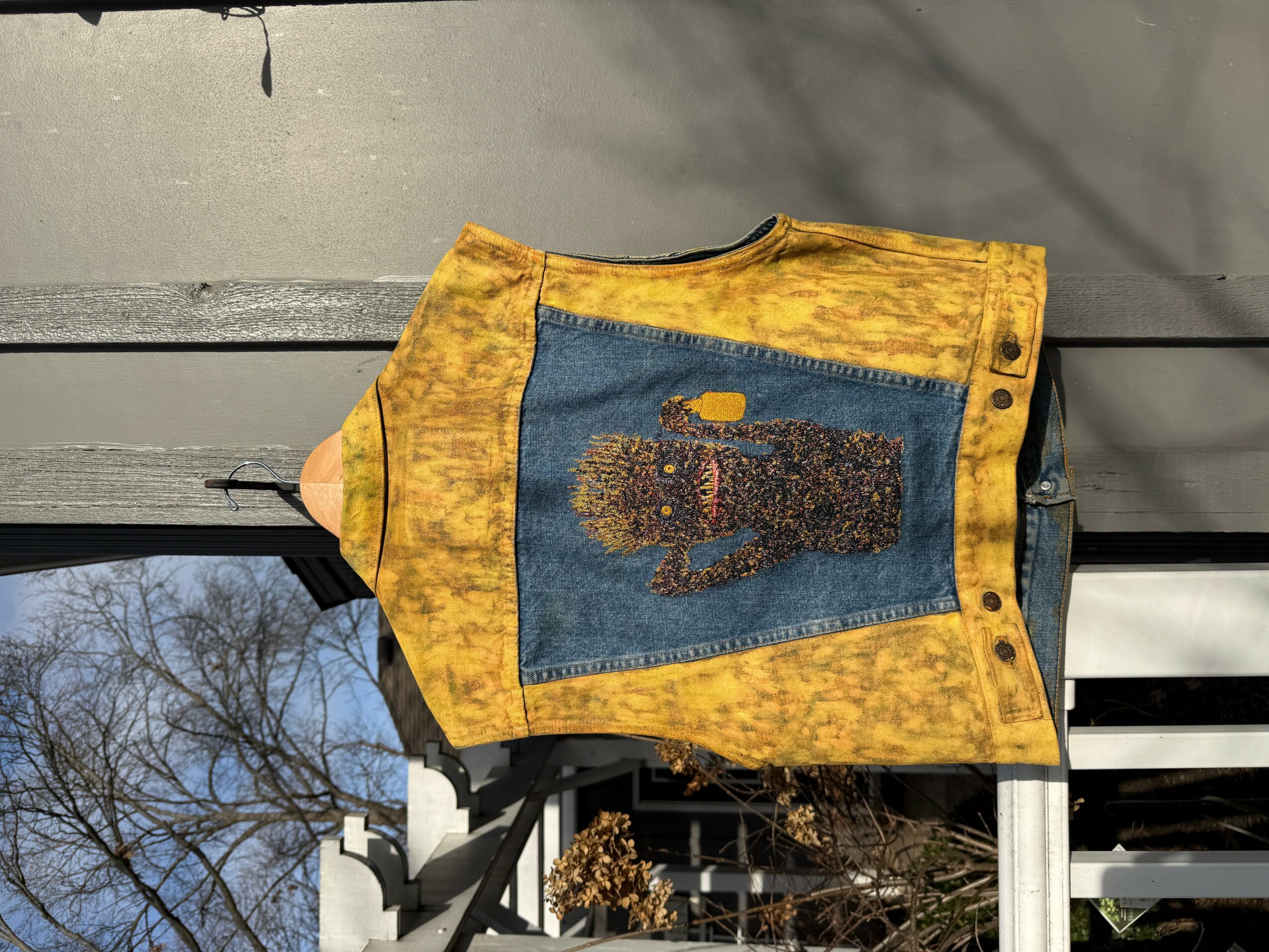 Yellow and blue vest with a bear design hanging outdoors, with trees and a building in the background.