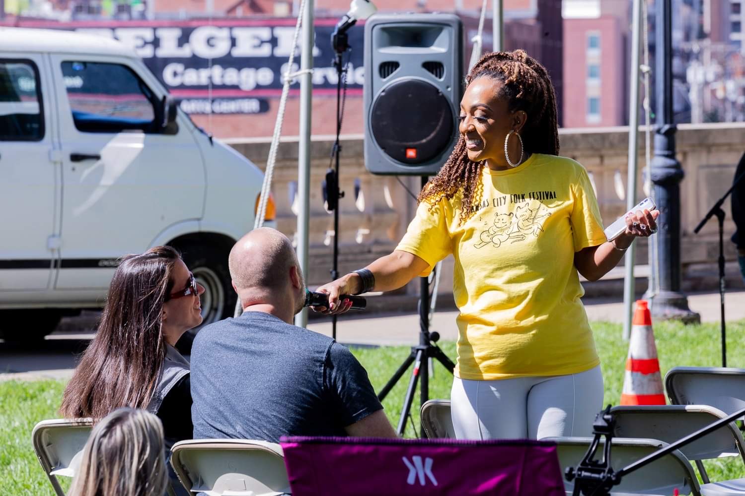Woman in yellow shirt singing into microphone outdoors, with three seated people listening, city background, speaker, and traffic cone.
