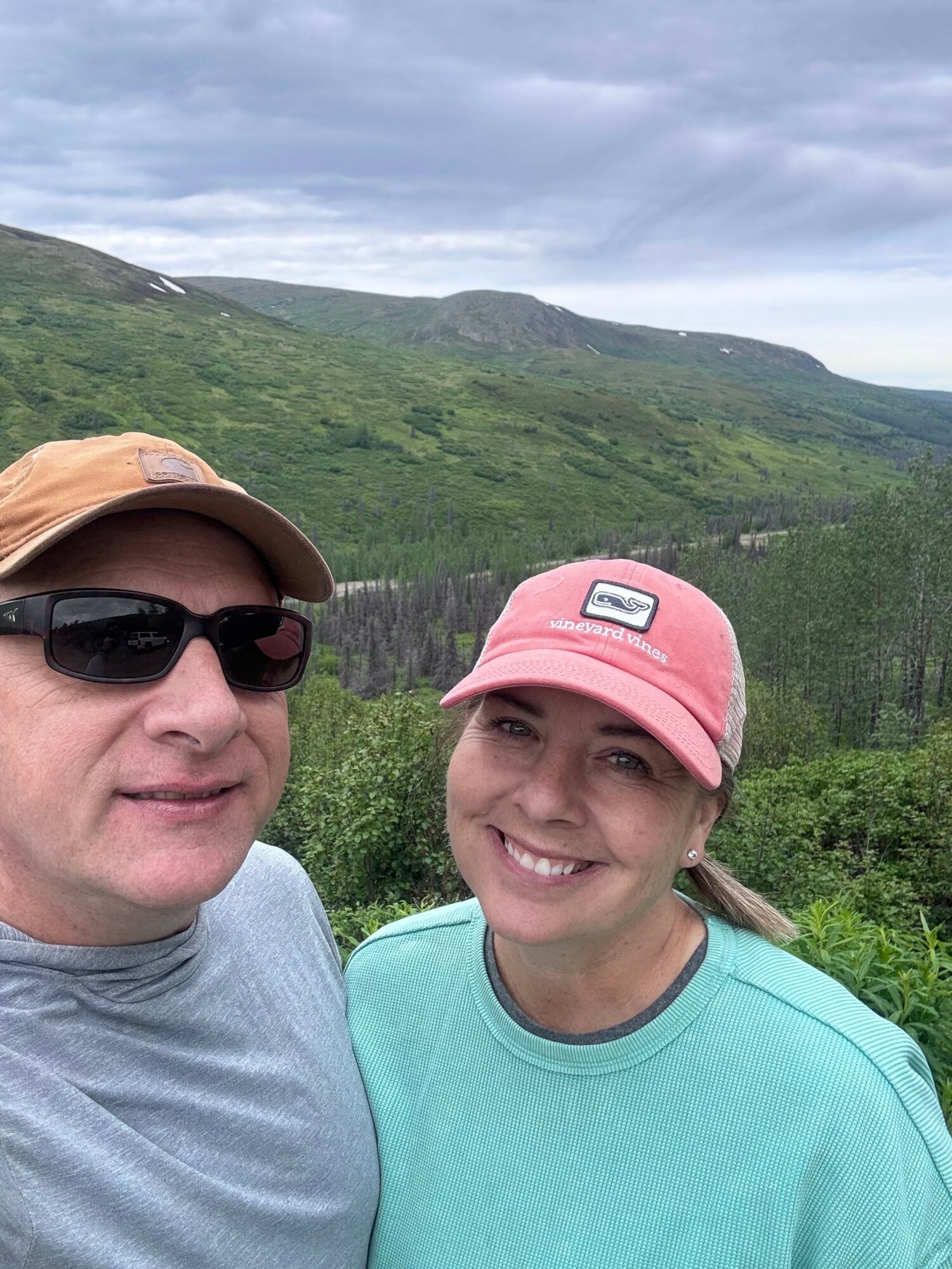 Two people outdoors with green hills and cloudy sky in background, smiling, one wearing a pink cap and sunglasses, the other a beige cap.