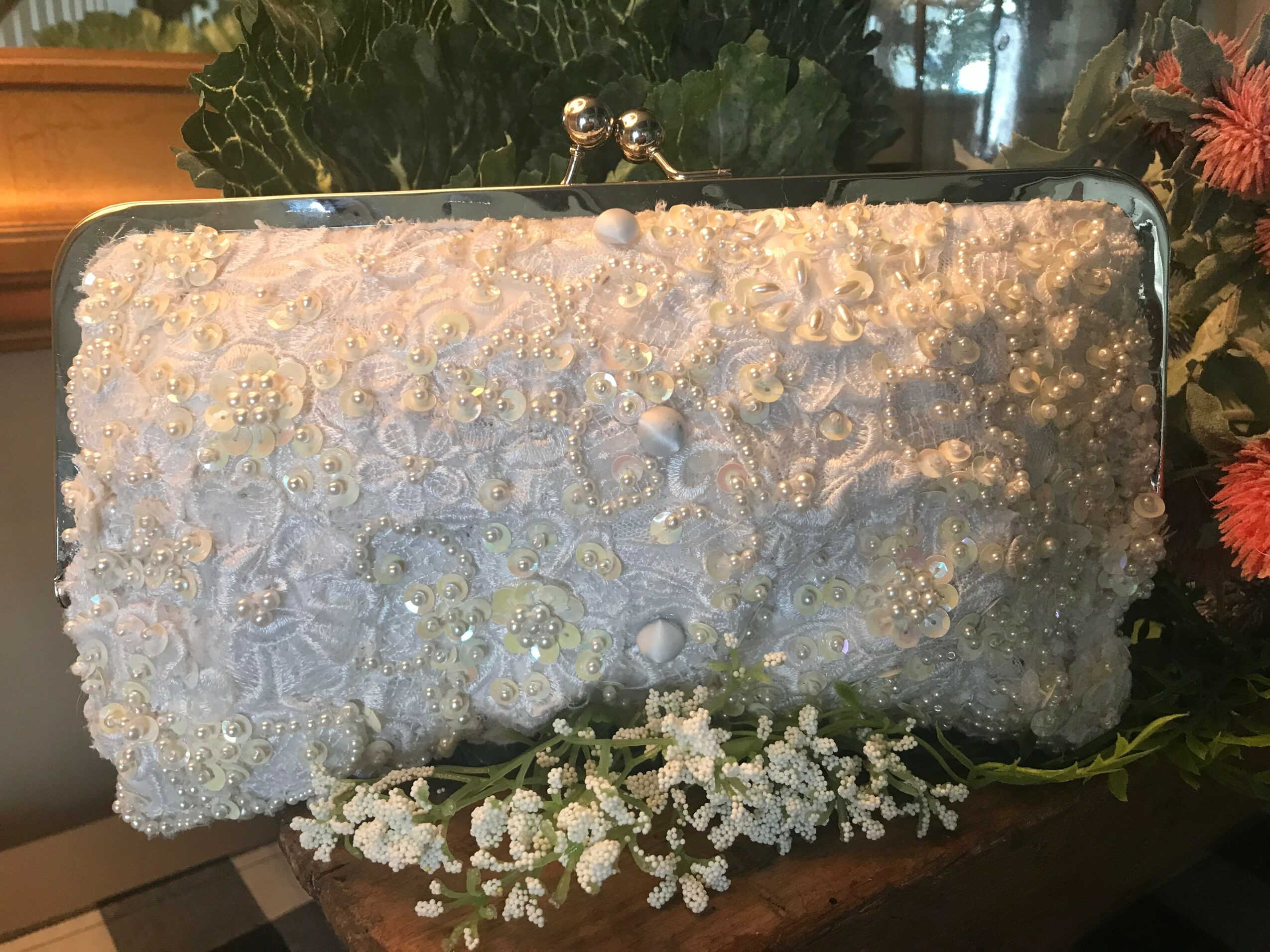 A rectangular white clutch purse with floral embellishments, placed on a surface with flowers and greenery around it.