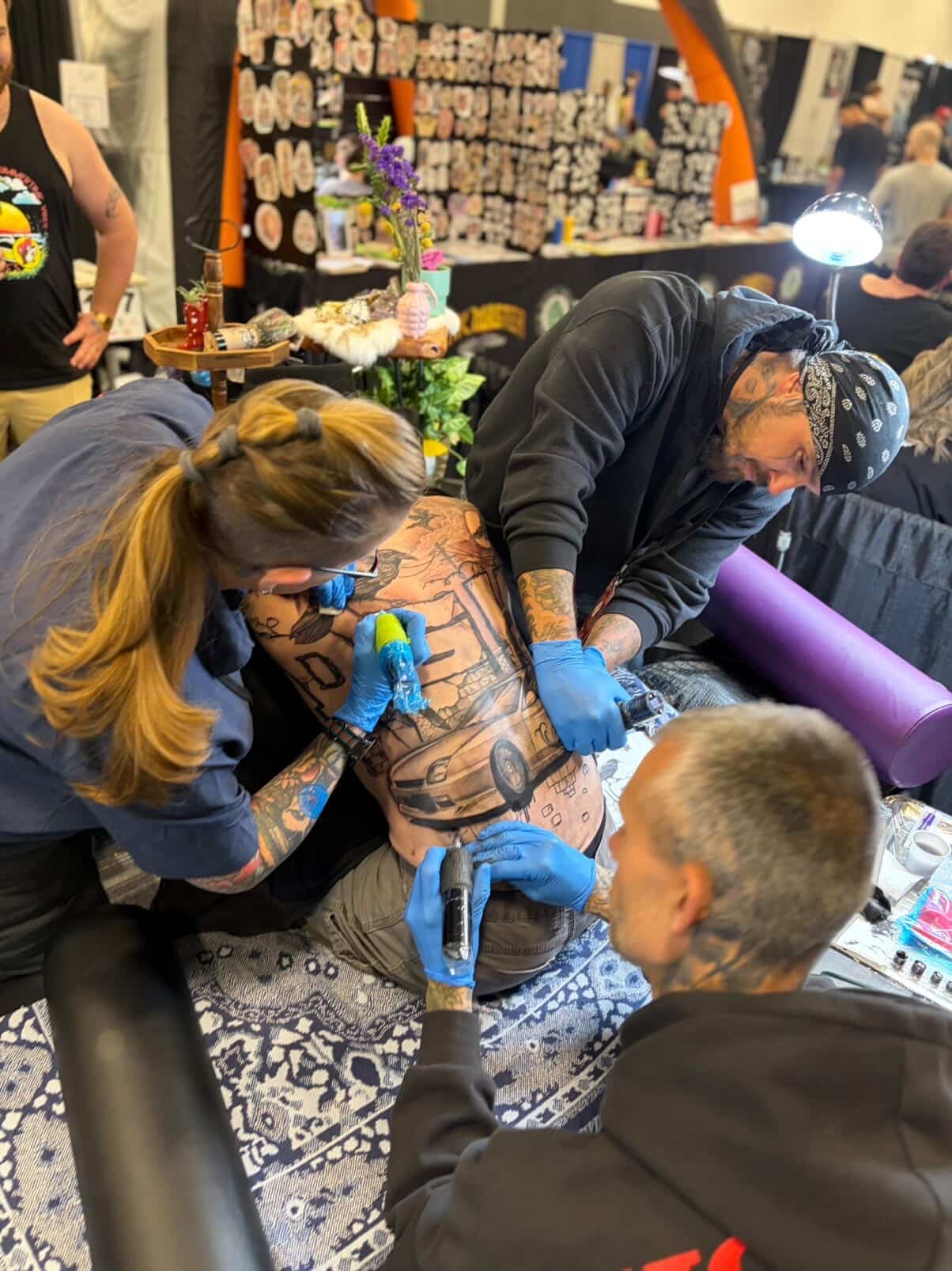 Tattoo artist working on a person's back in a busy shop with supplies and flowers nearby.