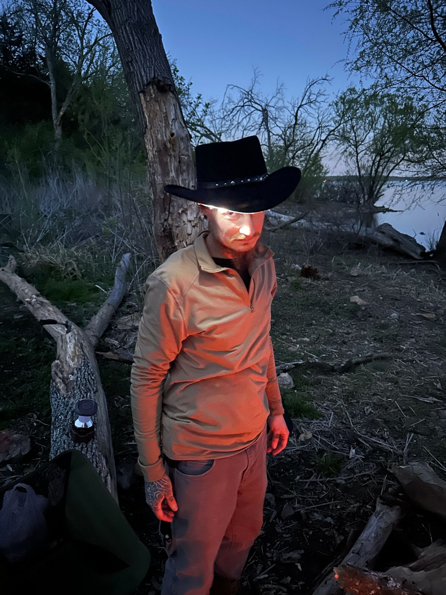 Person wearing a cowboy hat and orange jacket standing outdoors near trees and rocks.