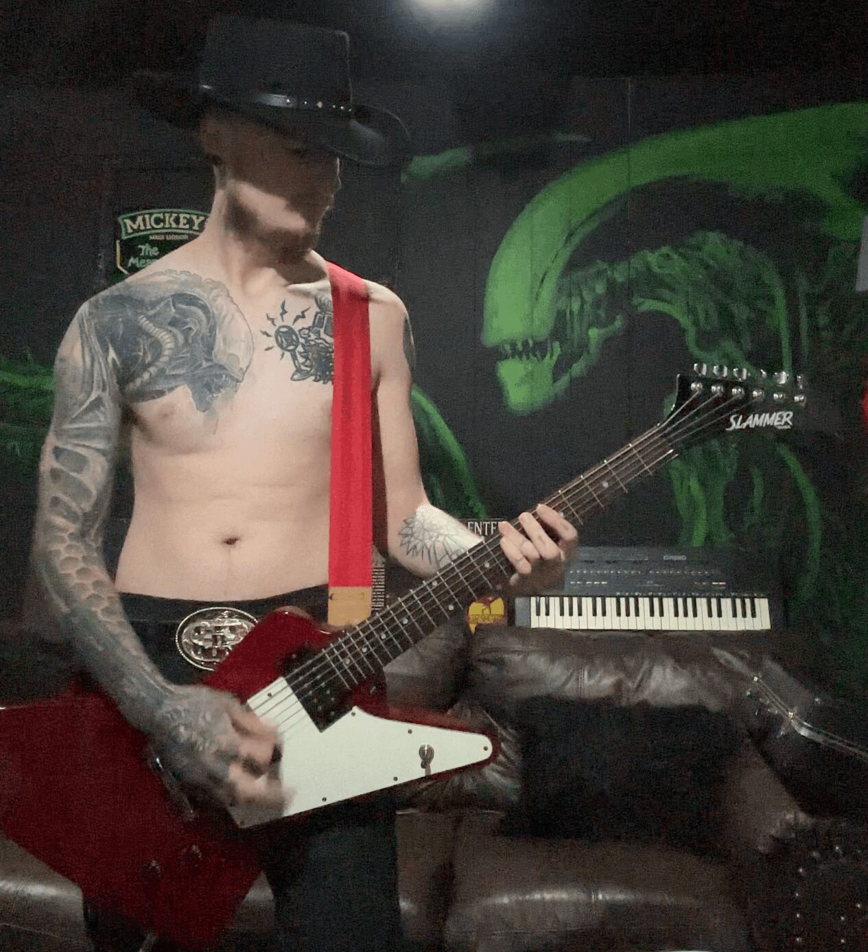 Tattooed shirtless person wearing a hat playing a red and white electric guitar in a dimly lit room.