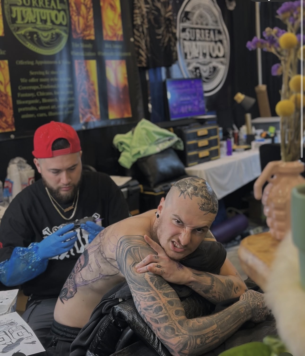 Tattooed man with short hair leaning on a table, looking at the camera, with a person in a red cap and blue glove behind him, in a tattoo shop.