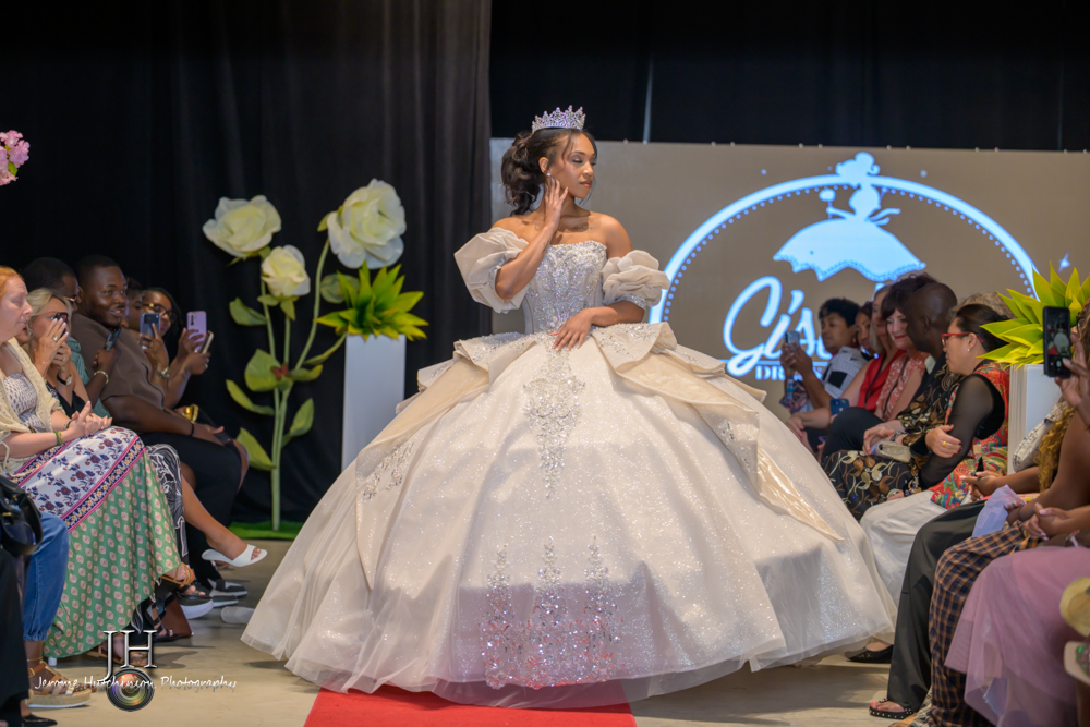 Woman in a wedding gown and tiara on stage, surrounded by seated audience, with large white flowers and a logo in the background.