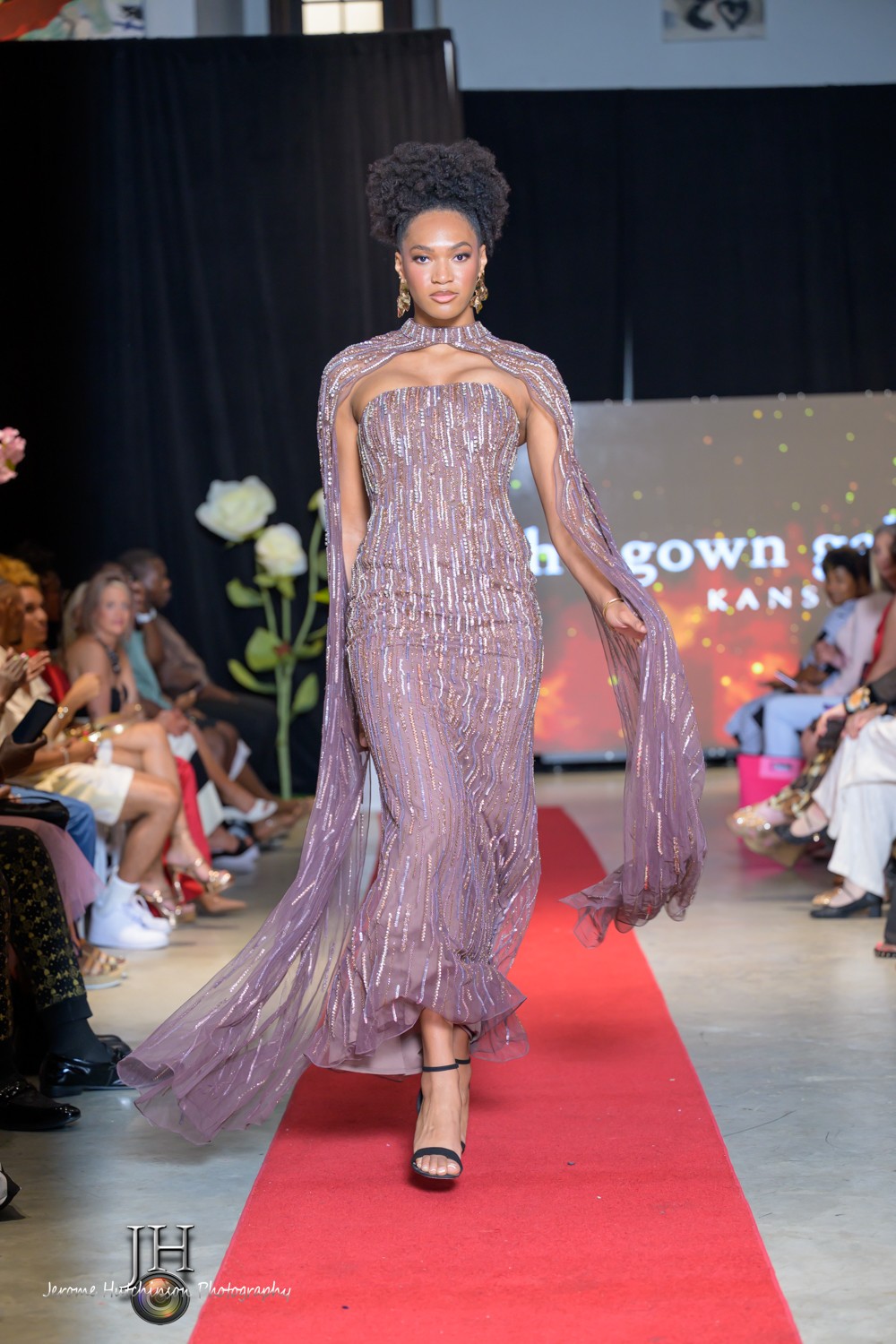 Model walking down runway in a shimmering, floor-length gown with sheer, flowing sleeves, surrounded by seated audience.
