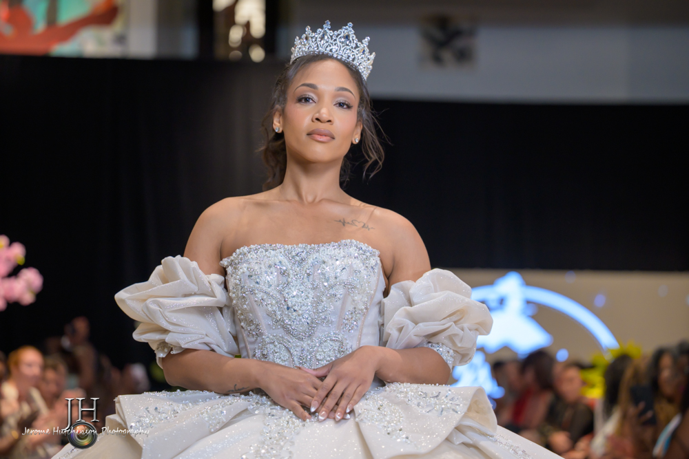 Woman wearing a crown and a silver gown with puffed sleeves, standing on stage with an audience in background.