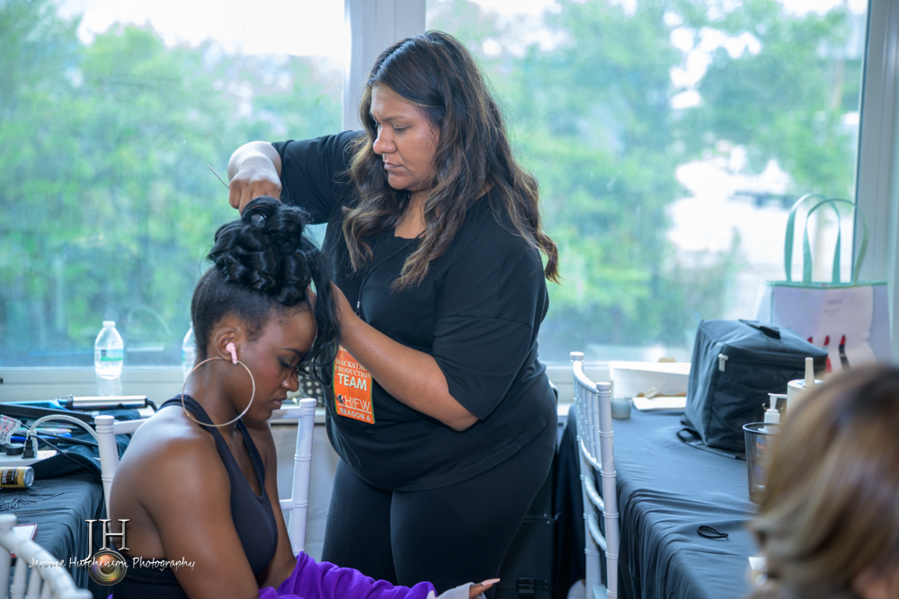 A woman styles a young woman's hair in a bright room with large windows and greenery outside.