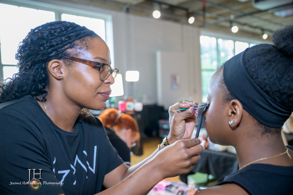 Makeup artist applying makeup to a woman with a black headband in a bright indoor space.