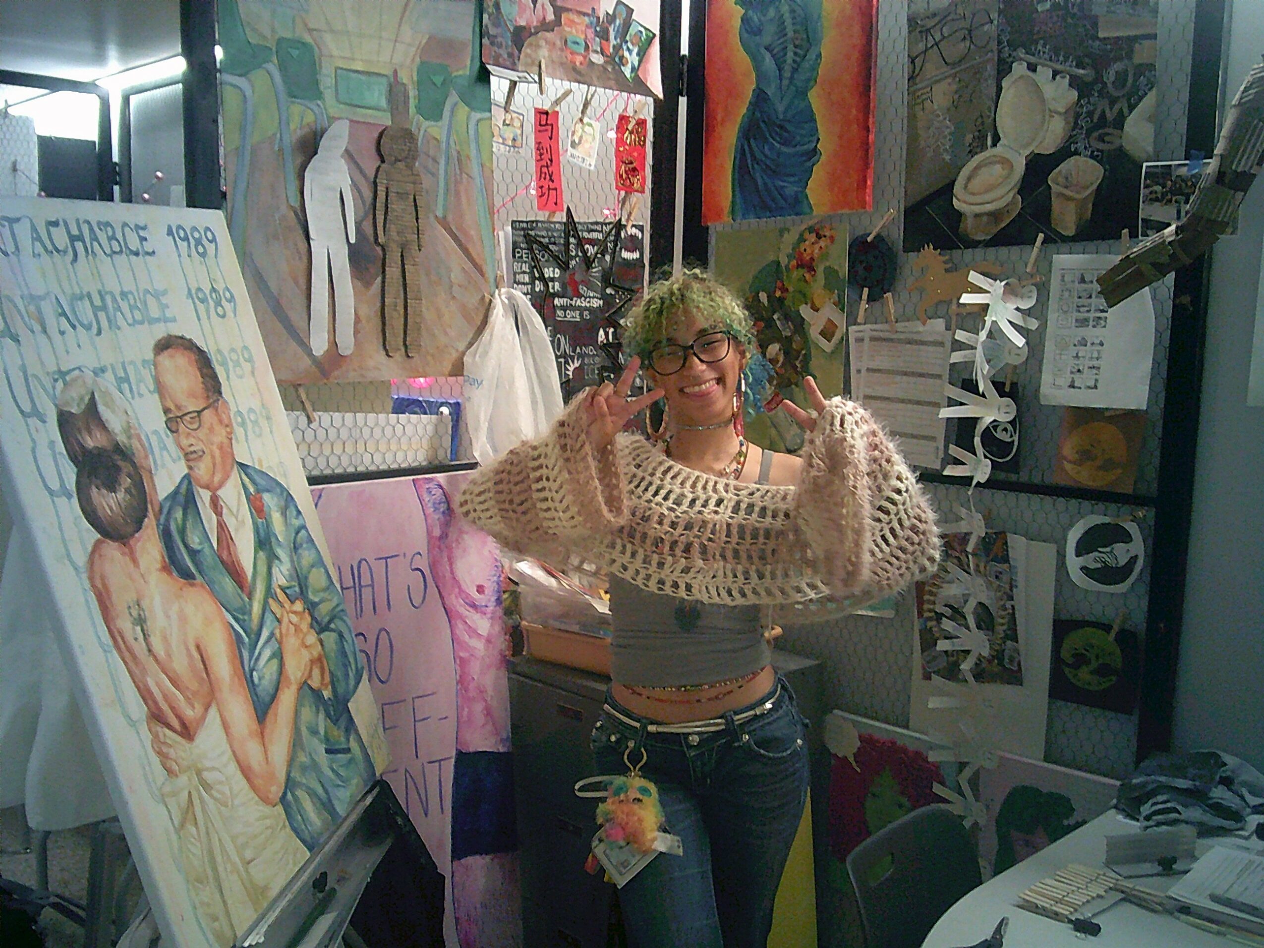 Person with glasses and curly hair smiling, taking a selfie in a room decorated with artwork and posters.