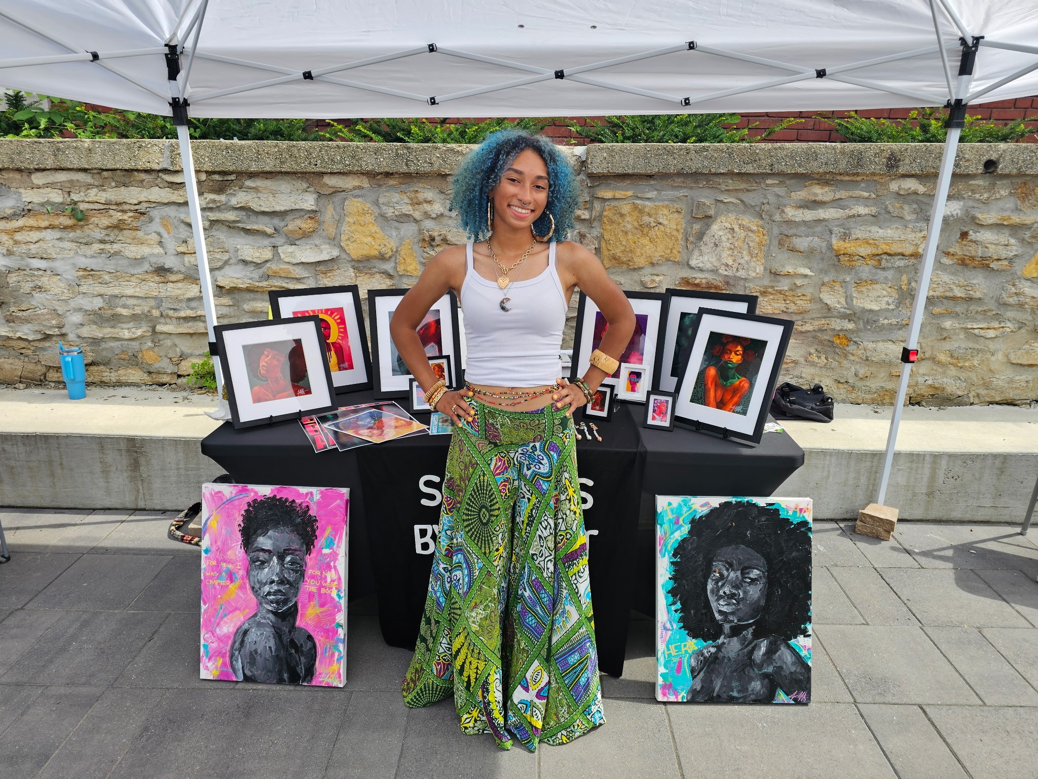 Young woman with blue curly hair standing outdoors, surrounded by framed artwork and paintings, under a white canopy.