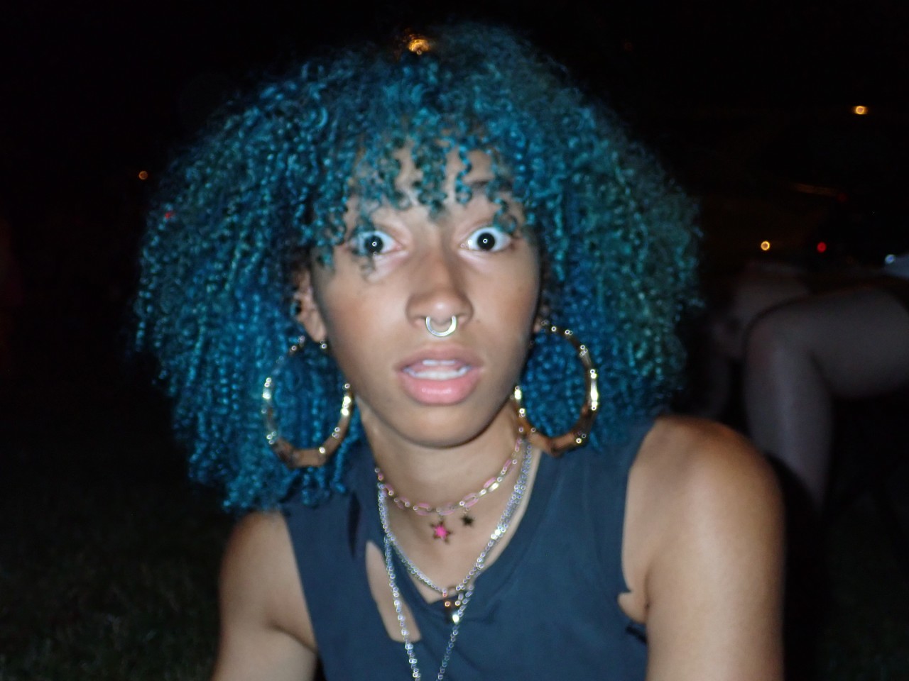 Person with blue curly hair, large hoop earrings, and multiple necklaces, looking surprised in a dark setting.