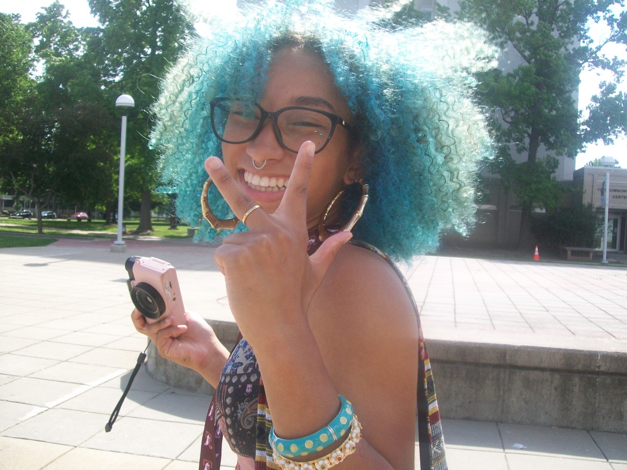 Smiling person with curly blue hair, glasses, holding a phone, making a peace sign, outdoors in a park.