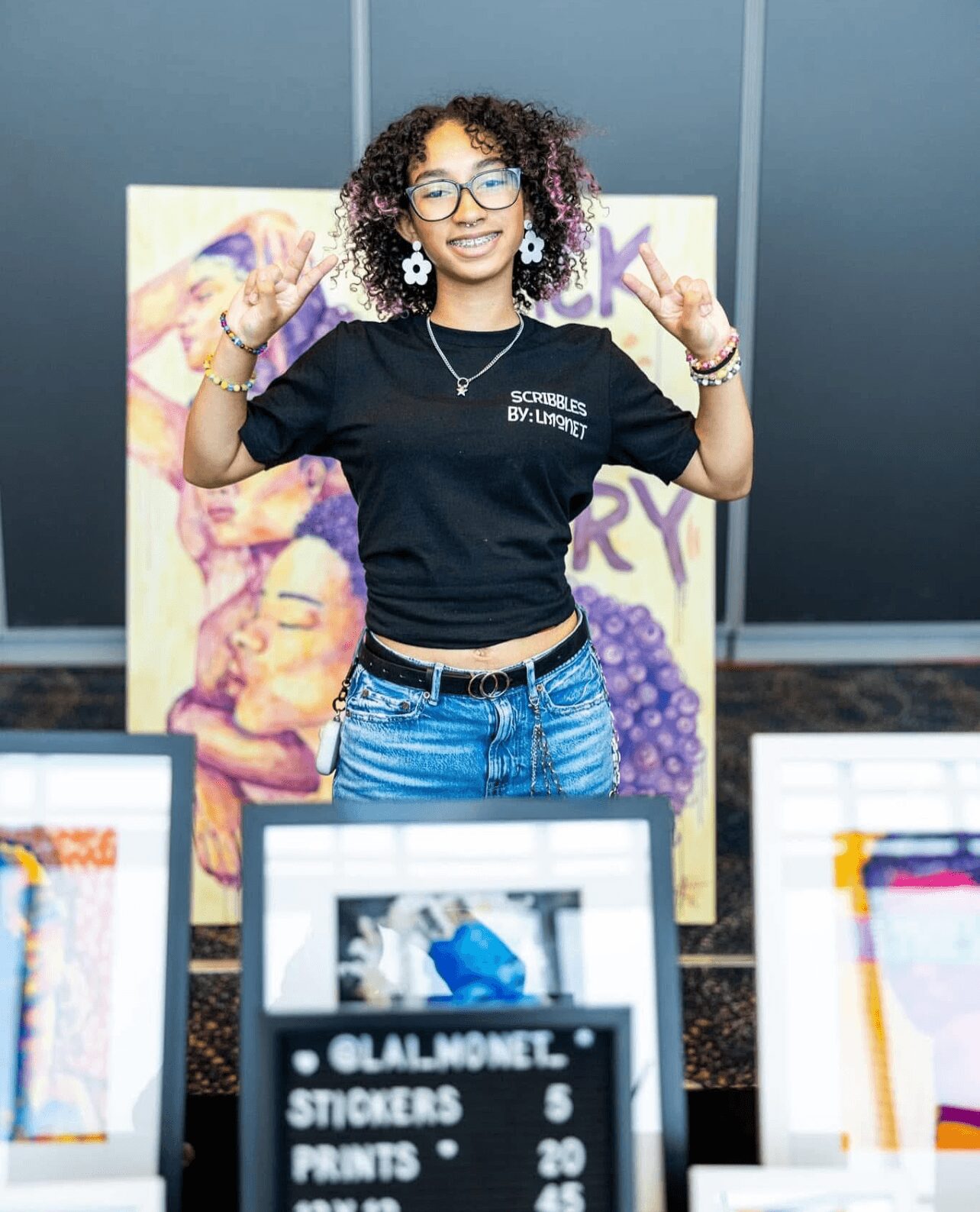 Young woman with curly hair and glasses smiling, standing in front of colorful artwork, wearing a black t-shirt and jeans.