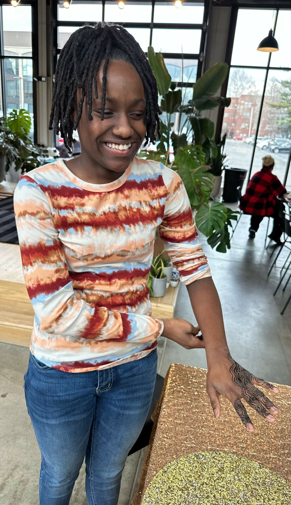 Smiling person with dreadlocks wearing a colorful striped shirt and jeans, standing indoors near a table.