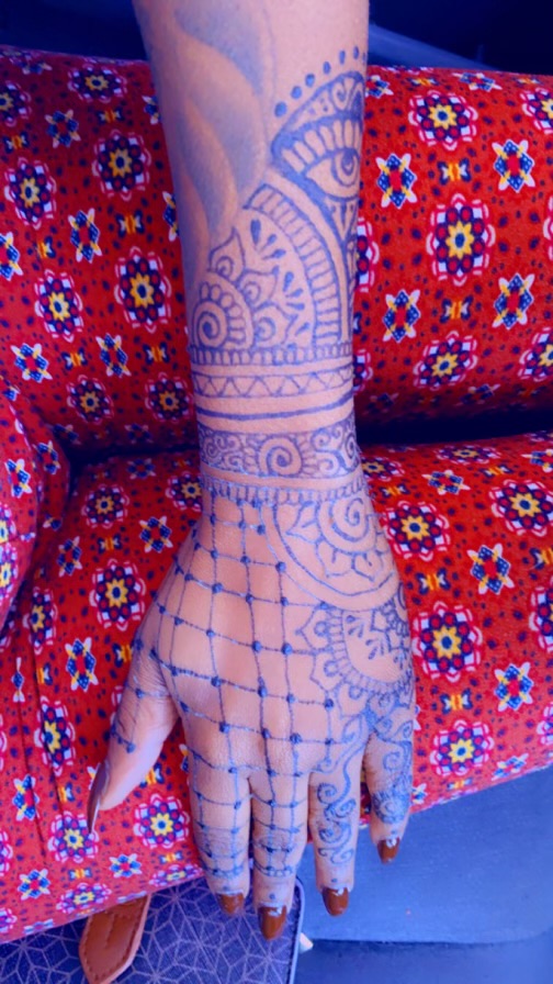 Hand and forearm with henna designs resting on a colorful patterned fabric, intricate patterns visible.