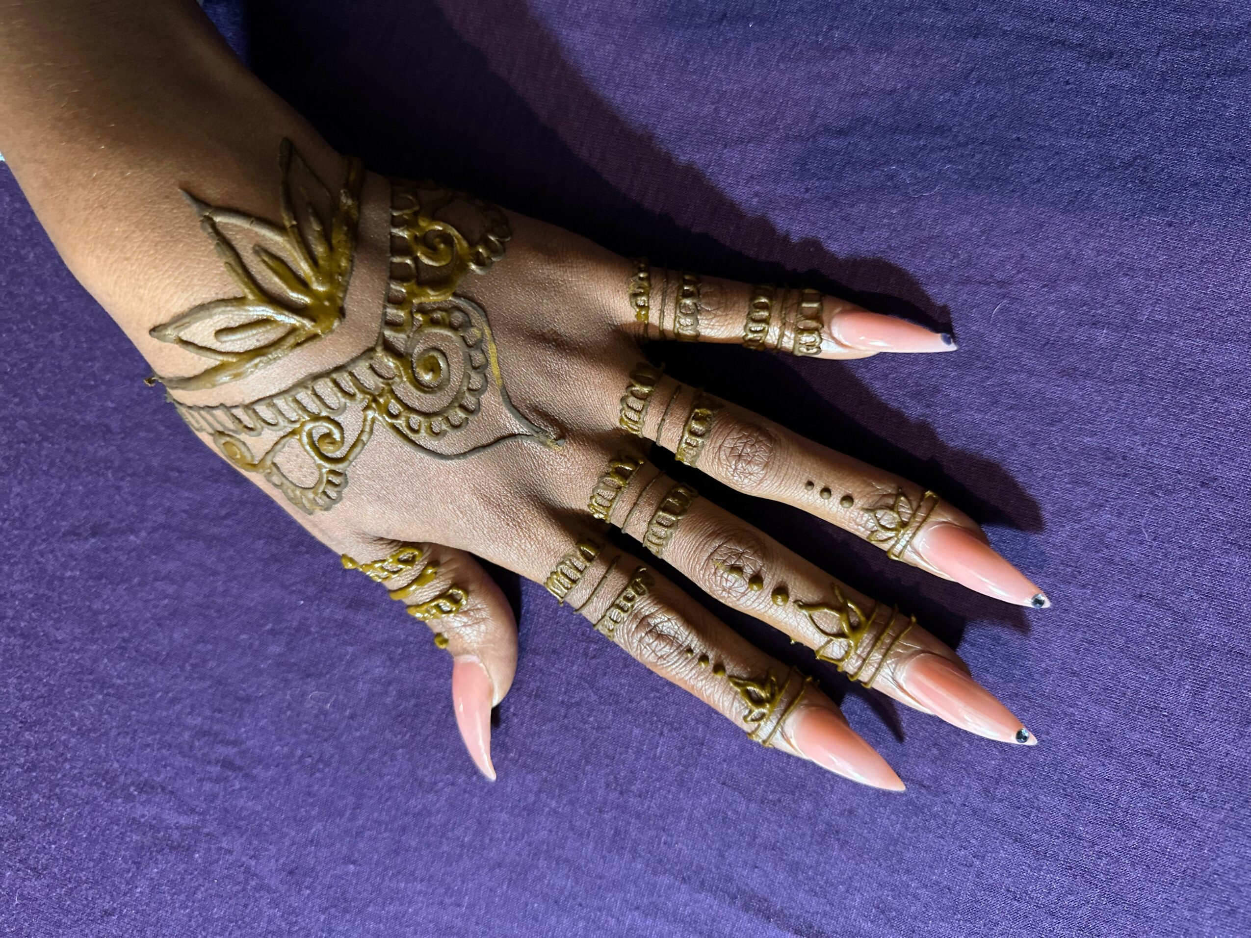 Hand with henna designs on fingers and back, resting on purple fabric, with long, pointed nails.