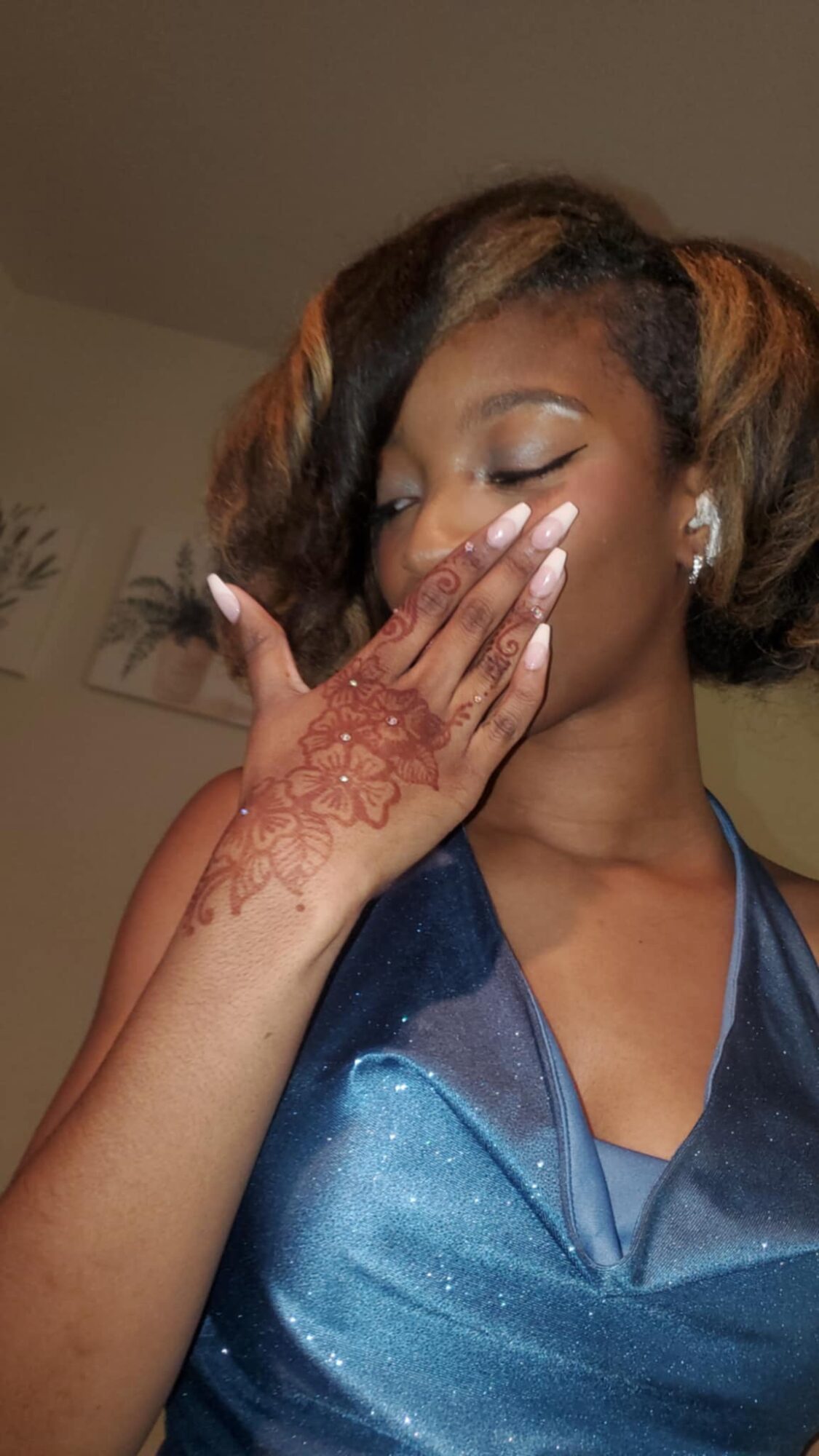 Young woman with styled hair, wearing a blue dress, covering her face with her hand showing henna on her arm.