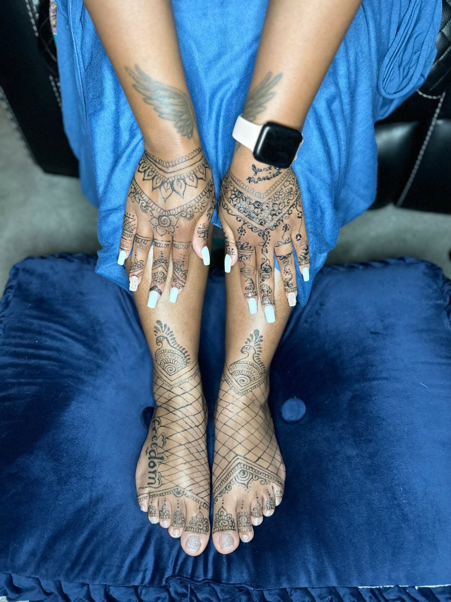 Person's hands and feet with intricate henna designs, wearing a smartwatch and blue clothing, resting on a blue surface.