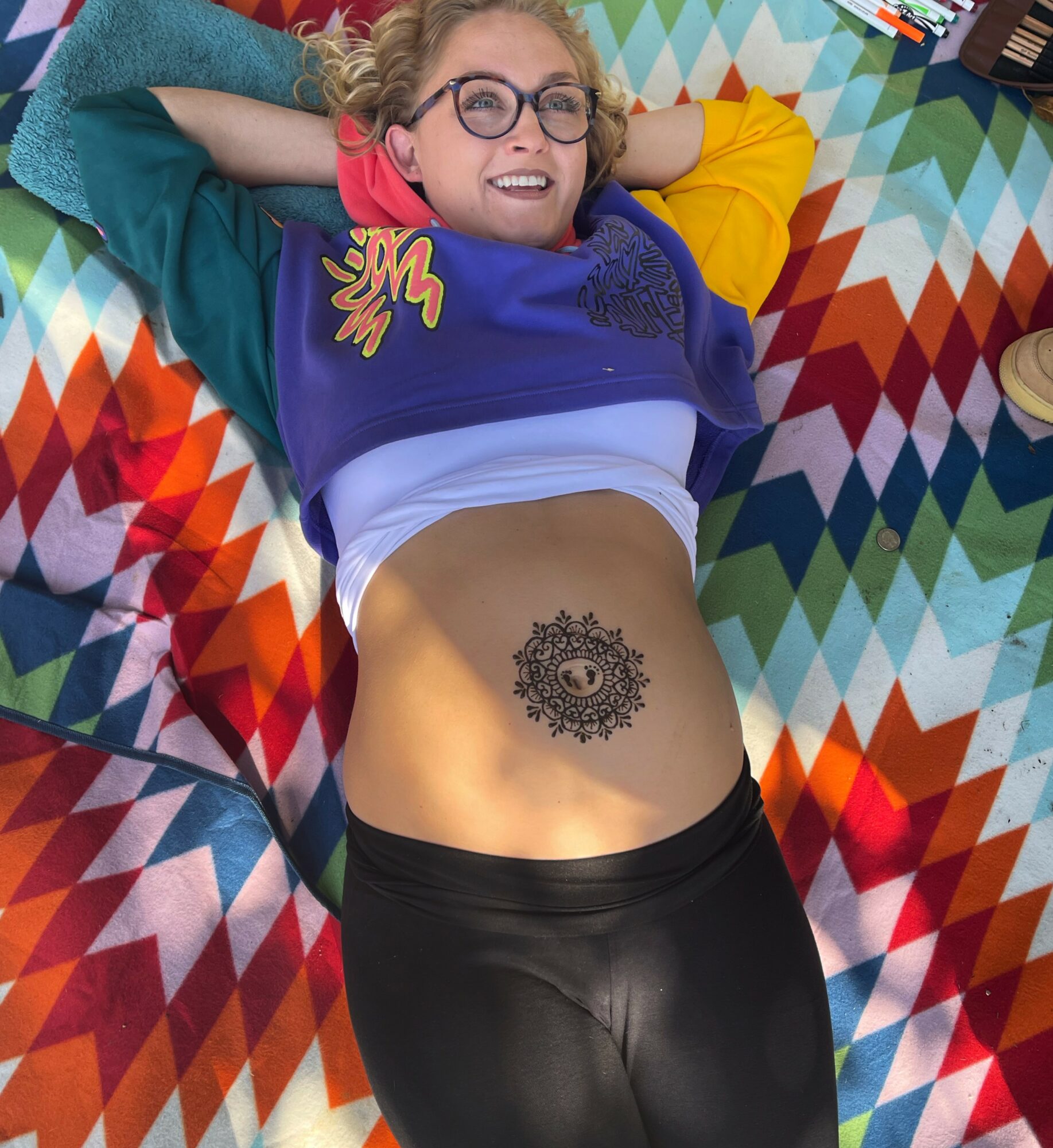 Young woman with glasses lying on colorful patterned blanket, smiling, showing her stomach with a decorative tattoo.