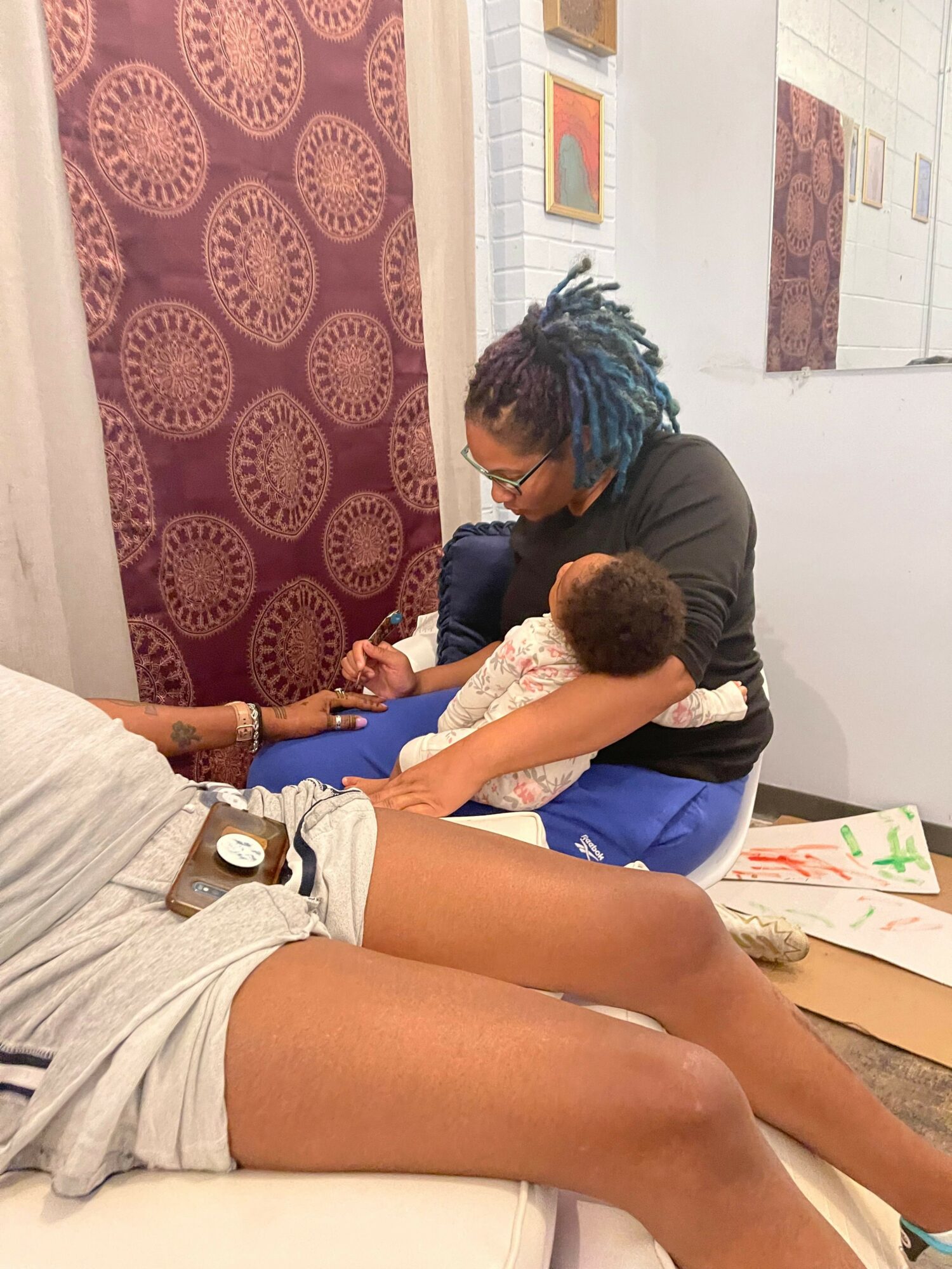 Person with blue and black hair holds a child while another person sits nearby, with legs extended on a white surface.