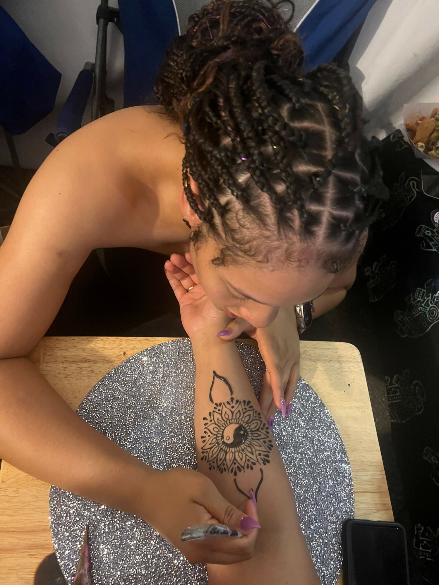 Person with braided hair signs a tattoo on their arm with a marker, sitting at a table with a glittery surface.