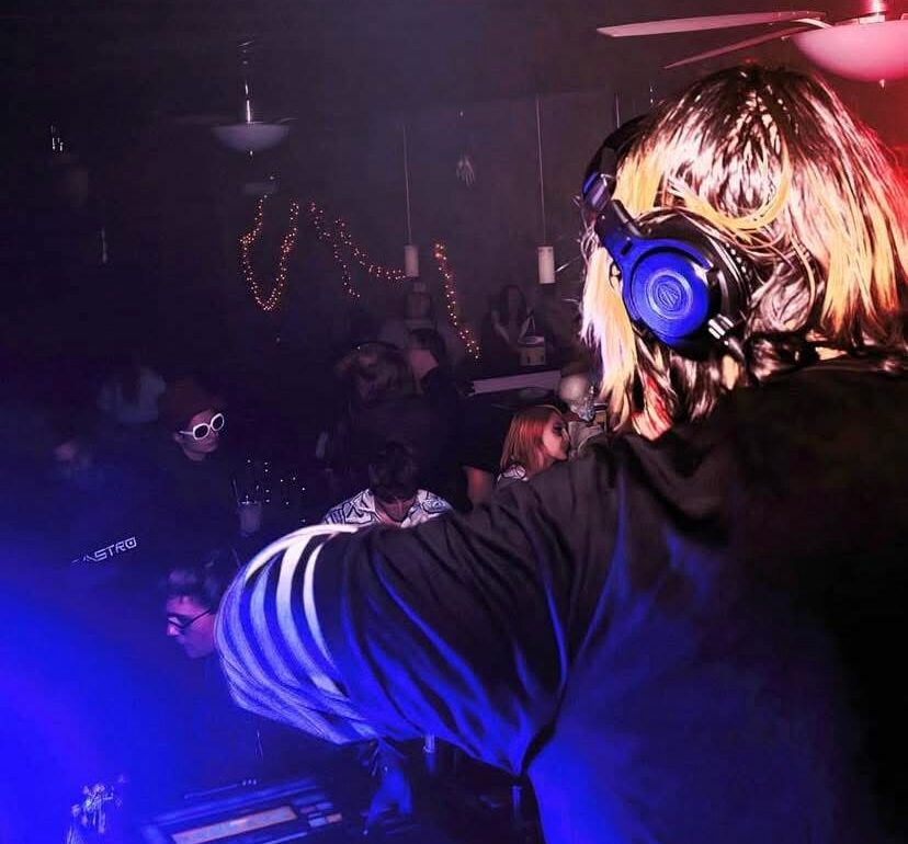 DJ with headphones performs at a nightclub with a crowd and colorful lighting.