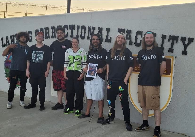 Group of seven people standing in front of a wall with text, some holding a framed photo, smiling.