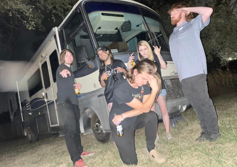 Group of five people standing and sitting outdoors near a bus at night, some making gestures, one with hand on head.