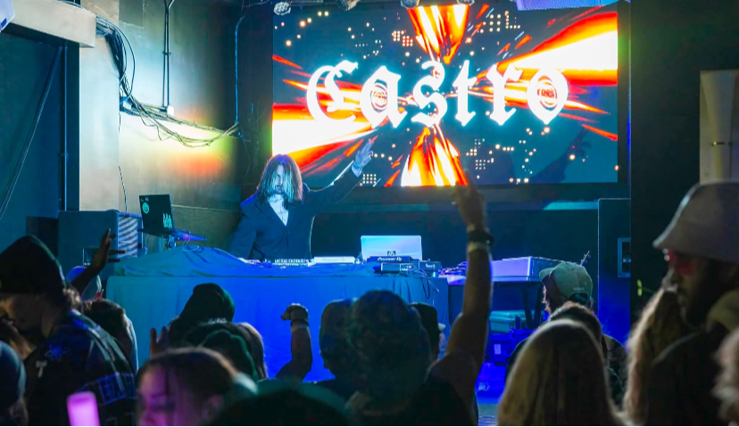 DJ performs at a club with a large colorful screen behind, crowd raising hands, and dim lighting.