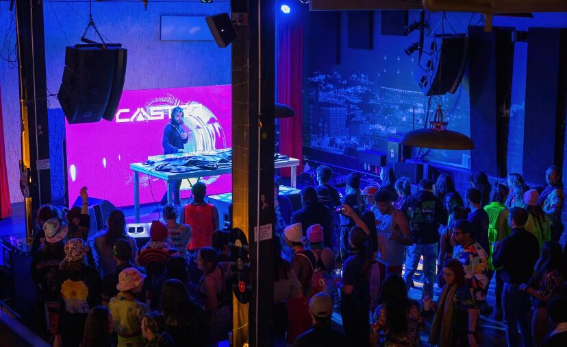 DJ performing on stage with a crowd in a dimly lit venue, blue and purple lighting, large screen behind DJ, audience watching.