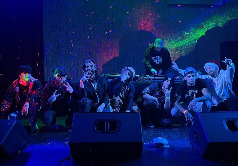 Group of seven people on stage with colorful background, some making peace signs, one person standing behind them, stage equipment visible.