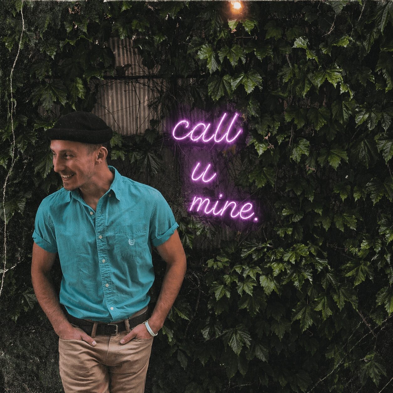 Person wearing a teal shirt and beige pants standing in front of a leafy wall with a neon sign that says 'call u mine.'