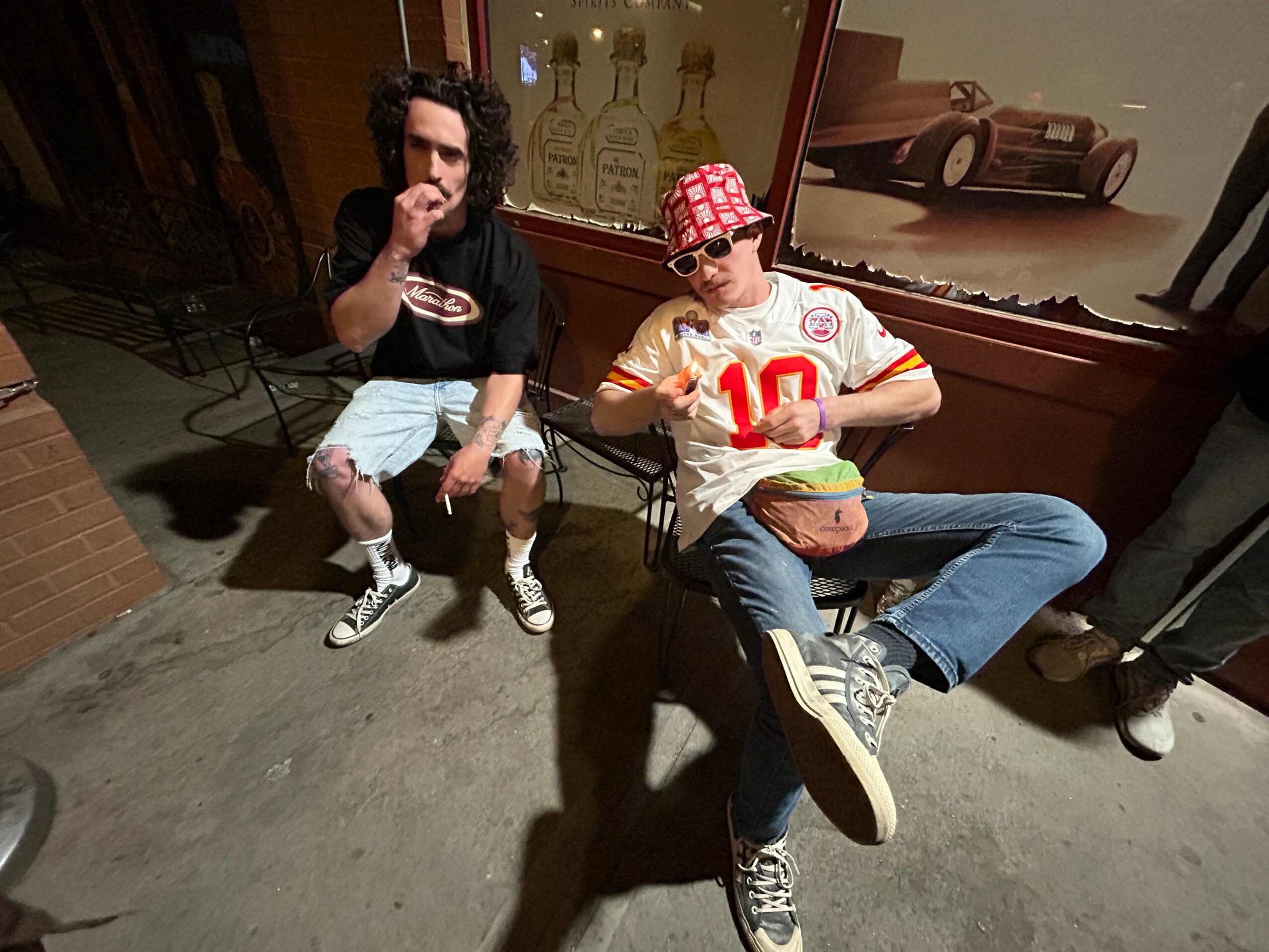 Two people sitting outside at night, one with curly hair and the other wearing a red checkered cap, holding a football.