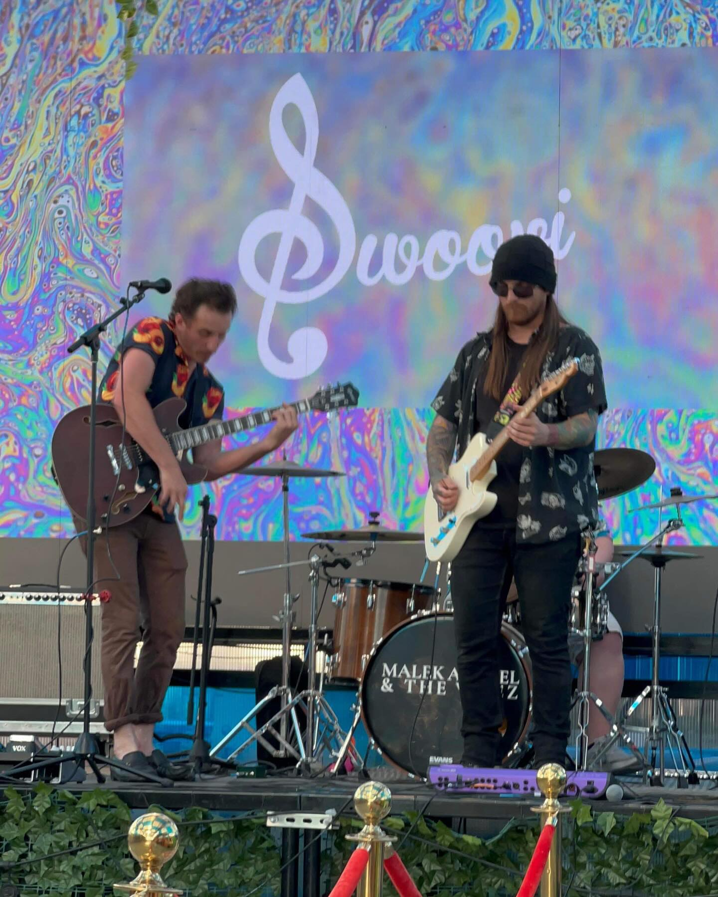Two musicians perform on stage with guitars, drums, and a colorful background screen displaying a treble clef and the word 'Swooi'.