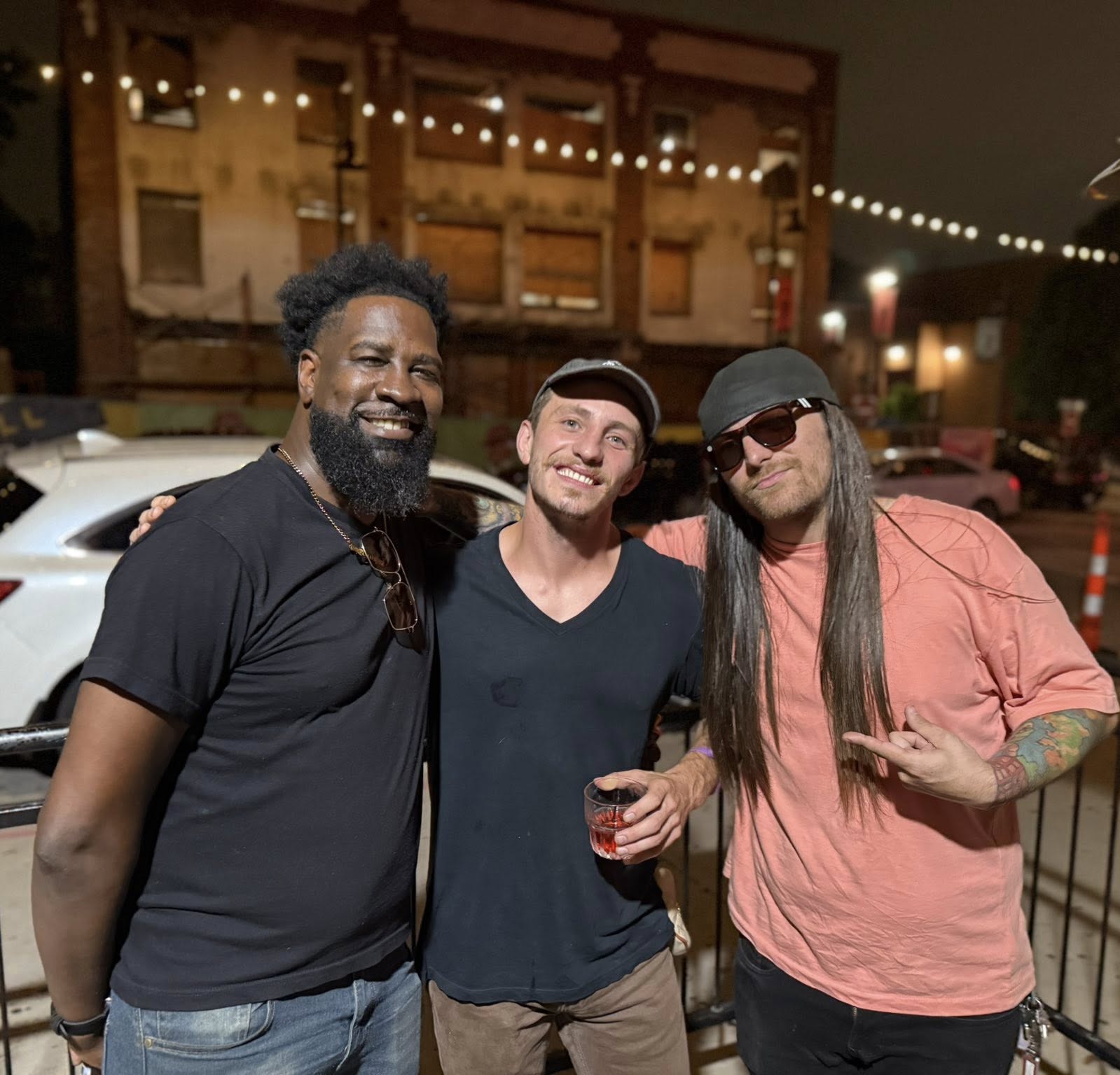Three men standing outdoors at night, smiling, with string lights overhead and buildings in the background.