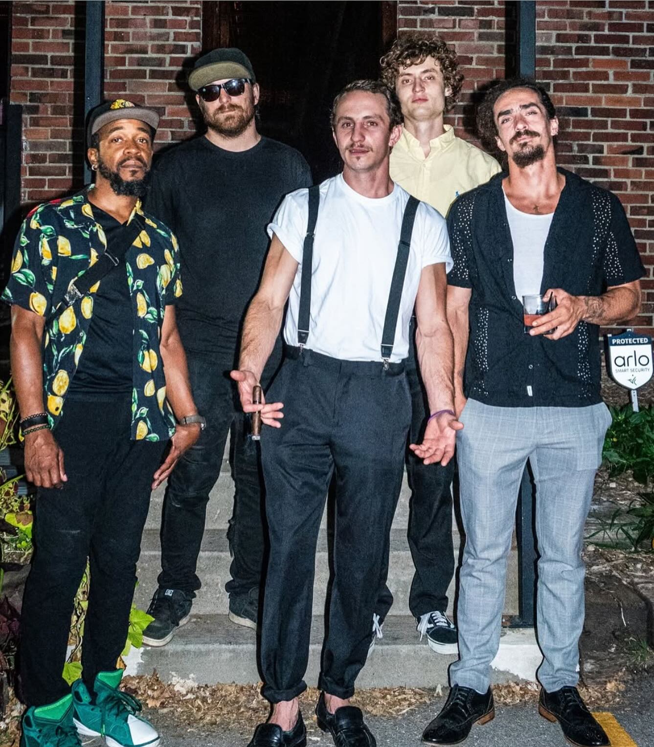 Group of five men standing outside in front of a brick building, dressed casually, some with drinks, smiling.