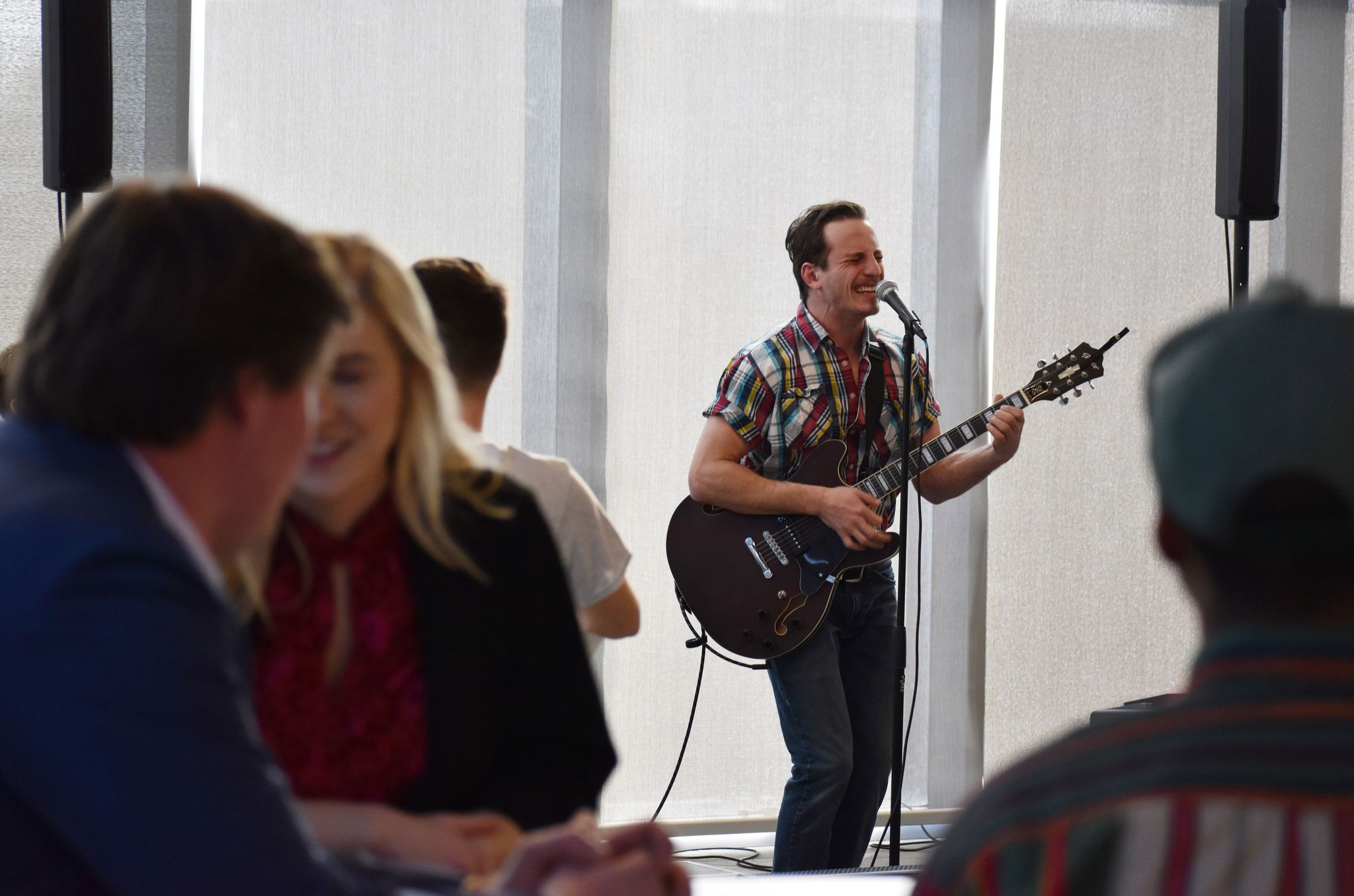 Man playing guitar and singing in front of a group of people indoors, with large windows behind him.