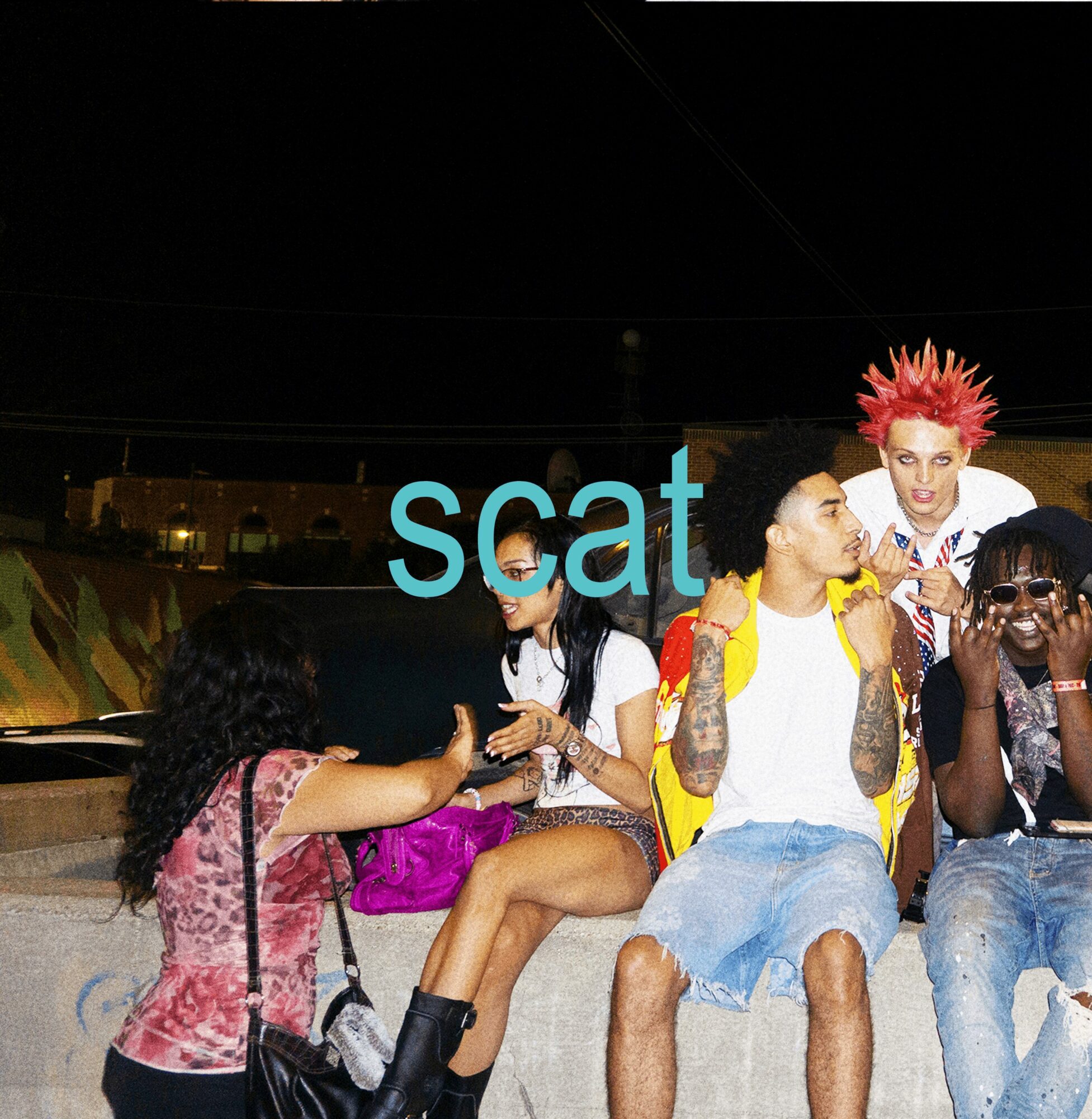 Group of young people sitting outdoors at night, some talking and others looking at their phones, with a partially visible word 'scat' in the background.