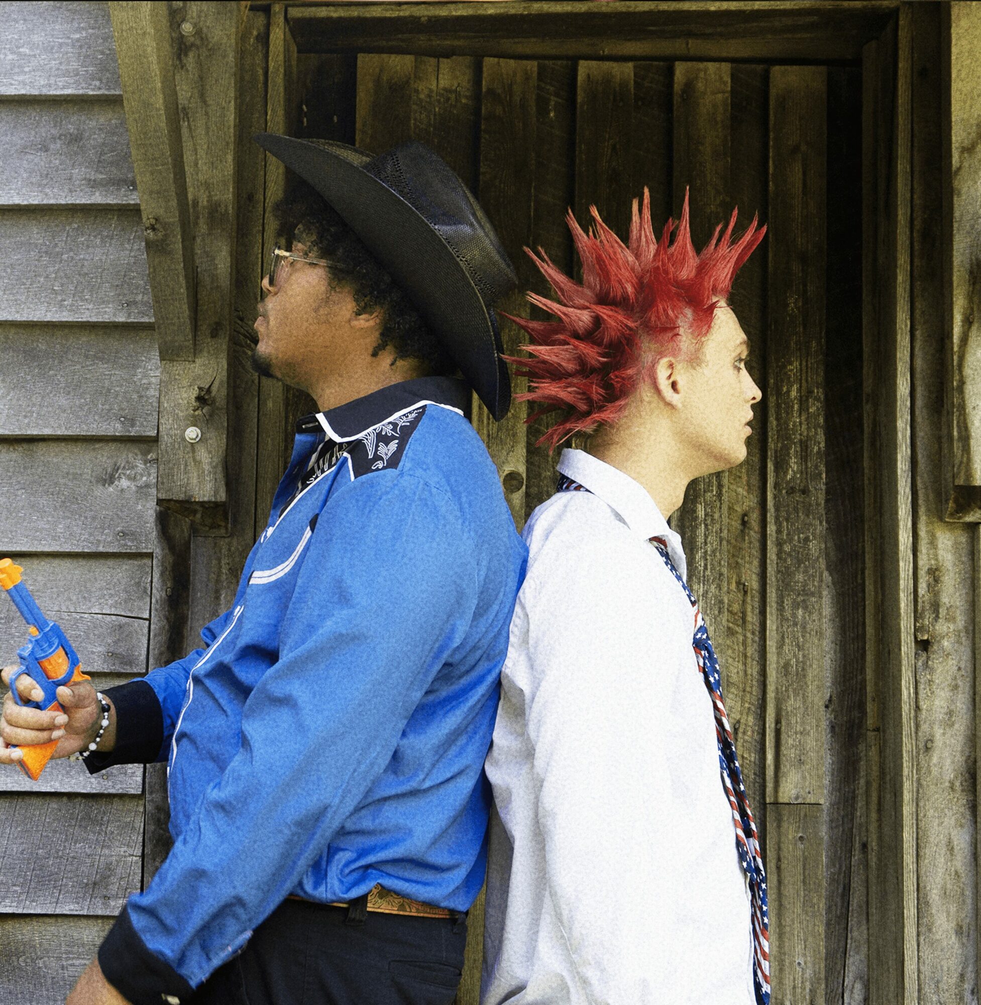 Two people stand back-to-back outdoors, one with a large hat and glasses, the other with spiked red hair, against a wooden background.