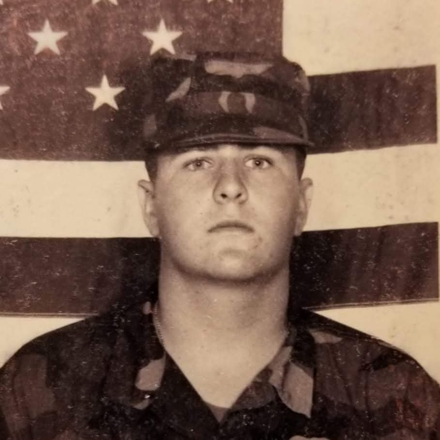 Young man in military uniform with a cap, standing in front of an American flag with stars and stripes.