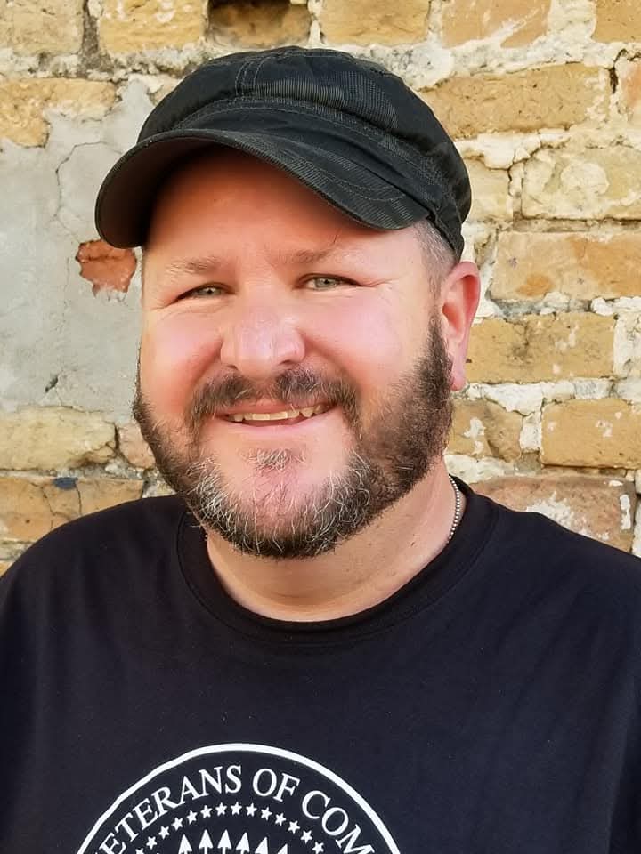 Man with beard wearing a black cap and black shirt, smiling, standing in front of a brick wall.