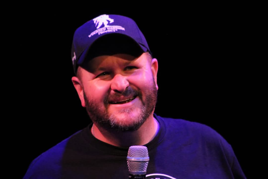 Man smiling, wearing a cap and holding a microphone, against a dark background.