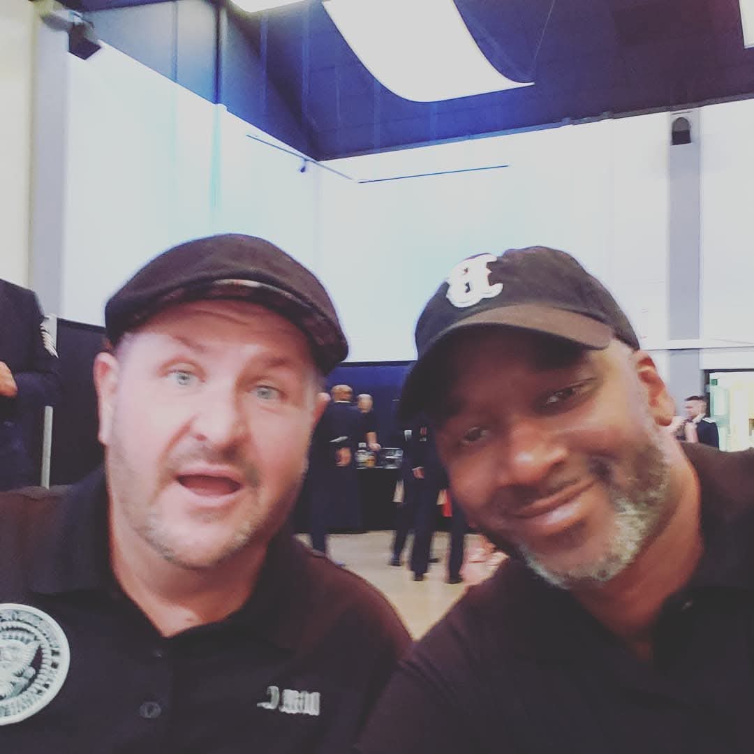 Two men smiling and taking a selfie indoors, with a high ceiling and bright lighting.