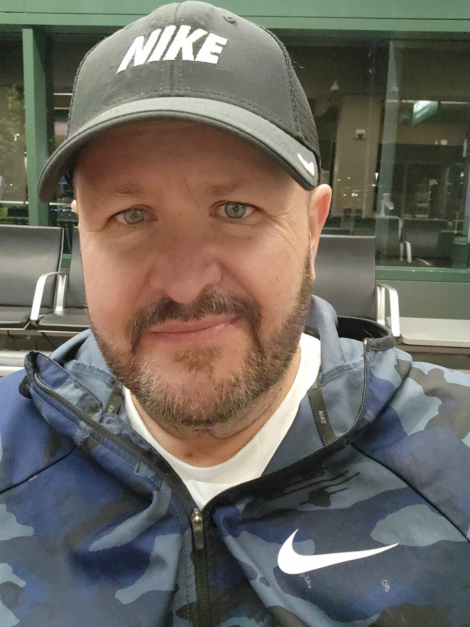 Man wearing a Nike cap and camouflage jacket, looking at the camera, with a building in the background.
